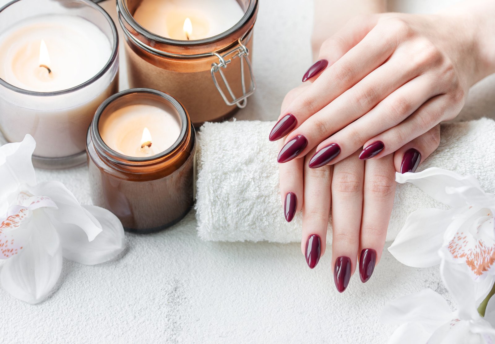 Beautiful hands of a young woman with dark red manicure on nails. Female hands on a towel in a nail salon, candles and white orchids flowers